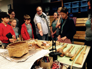 A man explaining the purpose of some artifacts laid out on a table to a group of school children