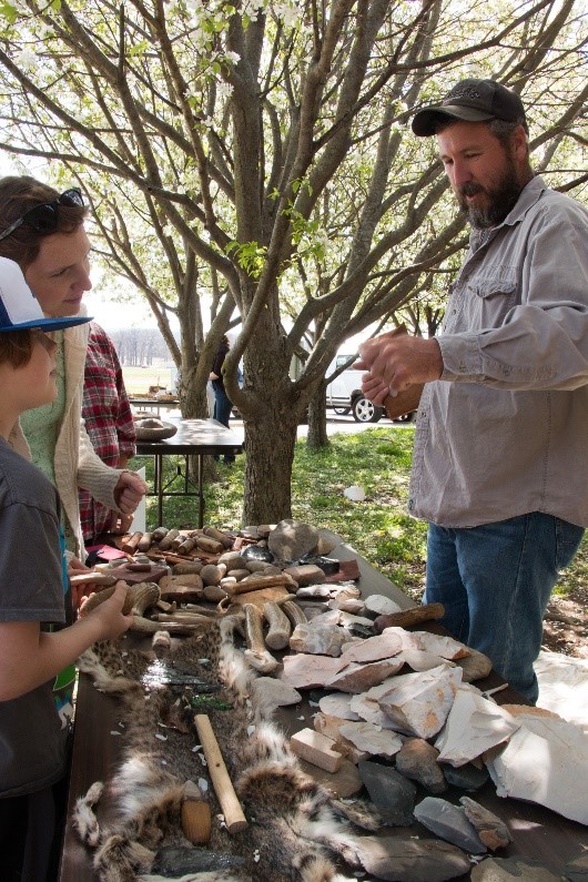 A tall, bearded man demonstrating replicas of Native American tools to a woman and her child under a shade tree.