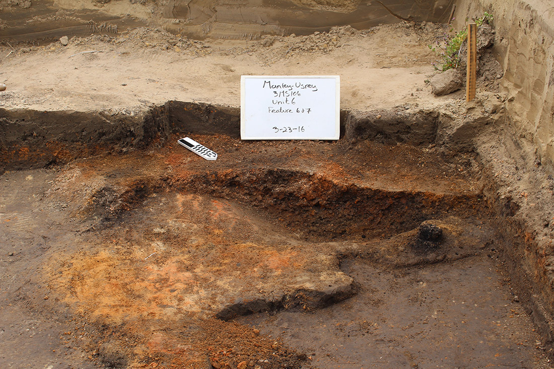 Figure 3: The hearth has been bisected and half of the fill removed.  Part of the floor was also burned and solid (orange area extending away from the bowl-like hearth).  An in situ wooden post was also found next to the hearth (black spot on the right near the edge of the hearth).