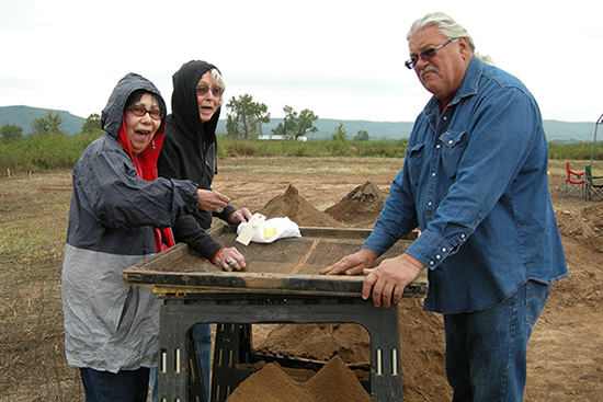 Three indigenous people sift excavation dirt through a wire screen.