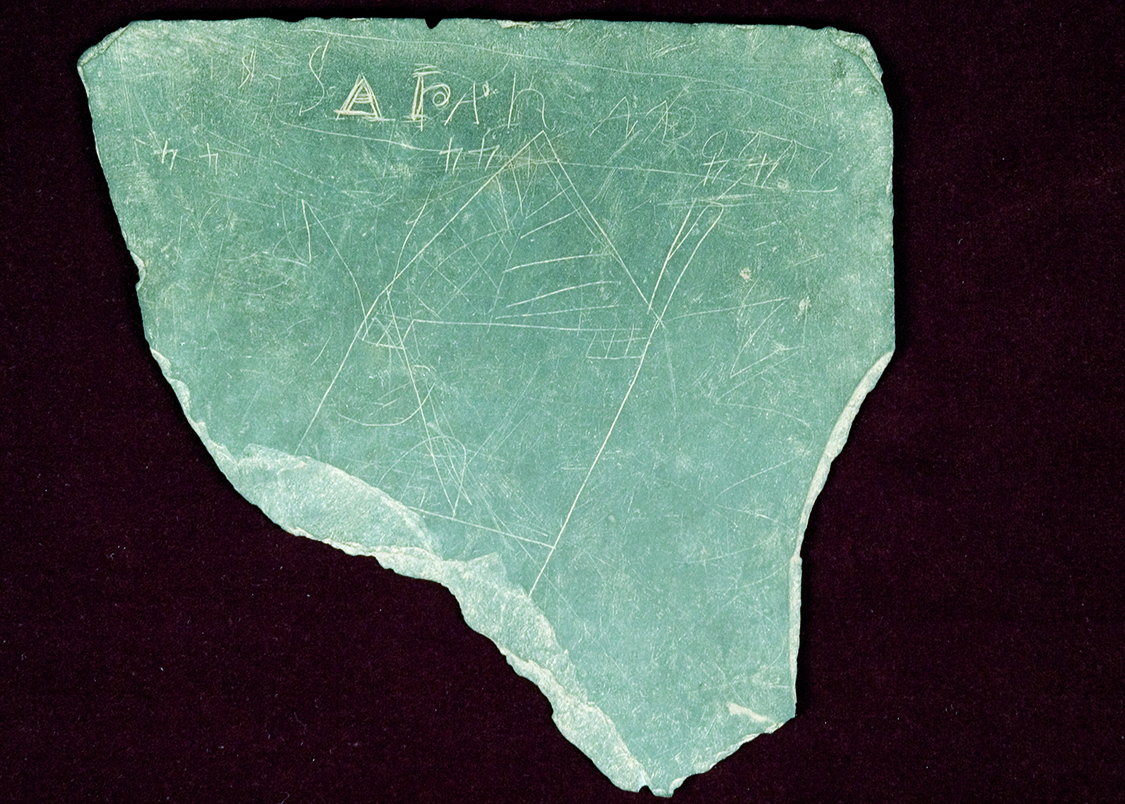 A writing slate fragment found in the Sanders House backyard during the 1981 excavations at Historic Washington Arkansas State Park. This slate is of particular interest because a picture of a house, the number “44” in three different locations, and the name “Sarah” have been permanently scratched into its surface.