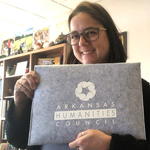 Photo of Mel Zabecki holding a gray and white Arkansas Humanities Council document folder.
