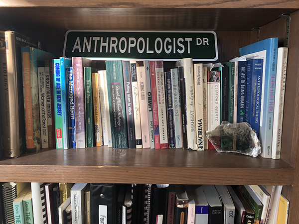 Photo of a dark wooden bookshelf with various anthropology and ethnography books and a rectangular green and white sign that says "Anthropologist Dr".