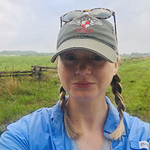 Photo of a young woman wearing a blue jacket and green ball cap standing in an open field.