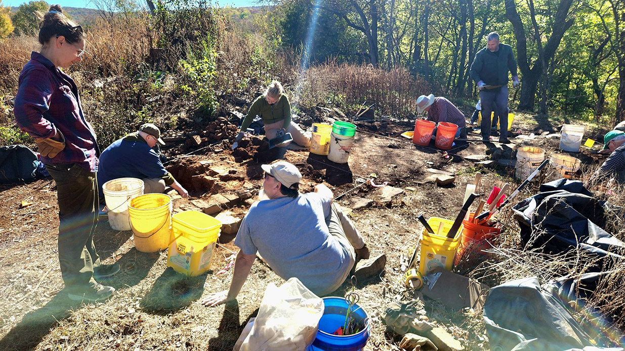 A group of people carefully excavating a stone structure foundation on a sunny spring day.