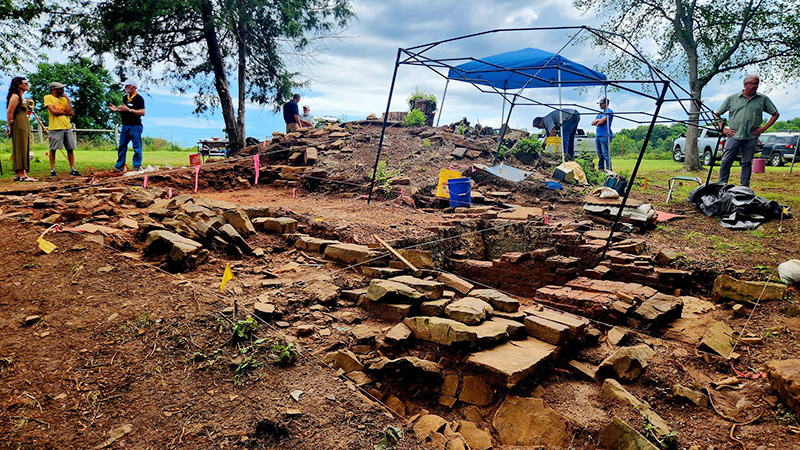 Photo of uncovered kiln foundation stones with archeologists and site visitors stand in the background.