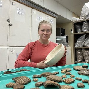 A woman wearing a red shirt holds a large pottery bowl in an artifact laboratory.