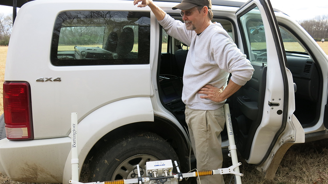 A man wearing a brown baseball cap and sunglasses stands next to a white SUV. 